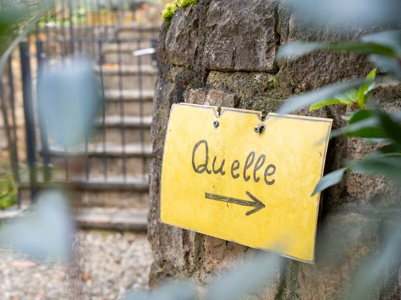 Ein gelbes Schild mit der Aufschrift "Quelle" und einem Pfeil nach rechts ist an einer Steinmauer befestigt, im Hintergrund sind Treppenstufen und ein Gitter zu sehen.