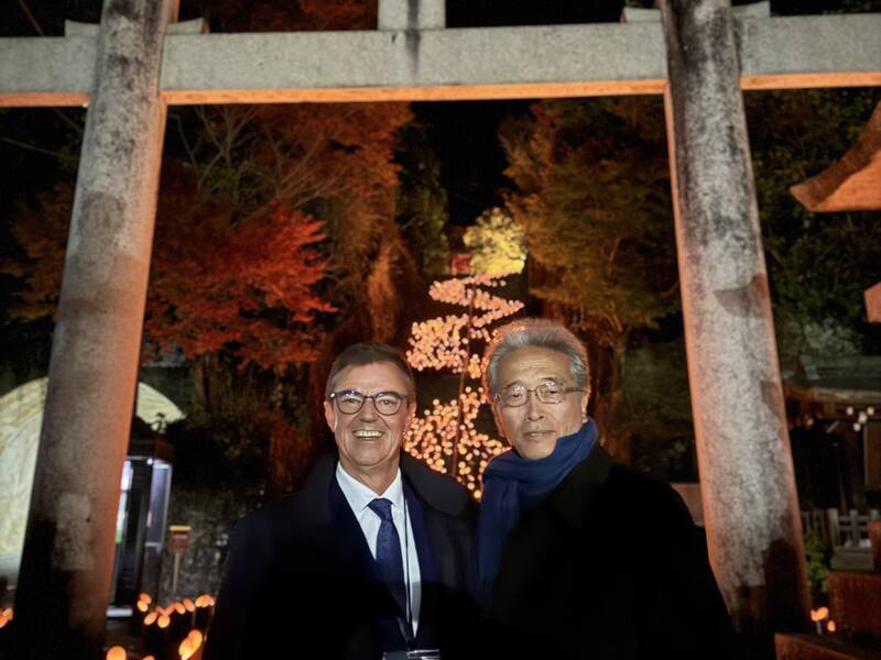 Zwei Männer stehen in einem nächtlichen, von einem Torii-Rahmen umgebenen Bereich, mit beleuchteten Bambusstäben und herbstlichen Bäumen im Hintergrund.