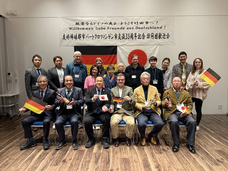 Eine Gruppe von Menschen sitzt und steht vor Bannern mit deutschen und japanischen Flaggen, einige von ihnen halten kleine Flaggen.