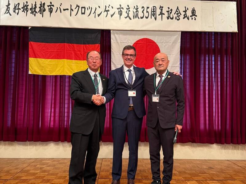 Drei Männer in Anzügen stehen auf einer Bühne vor den Flaggen Deutschlands und Japans und einem japanischen Banner, während sie sich die Hände schütteln.