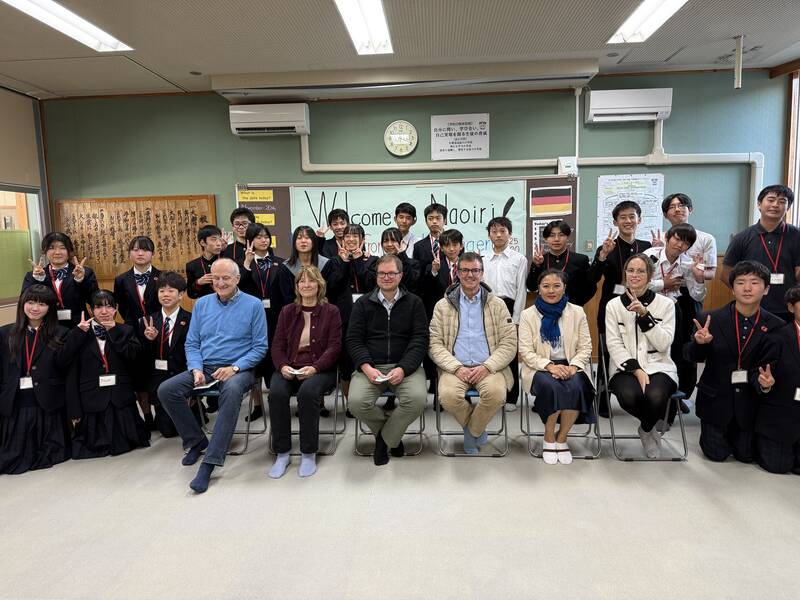 Eine Gruppe von Schülern und Erwachsenen posiert lächelnd für ein Gruppenfoto in einem Klassenzimmer mit einem Willkommensschild im Hintergrund.