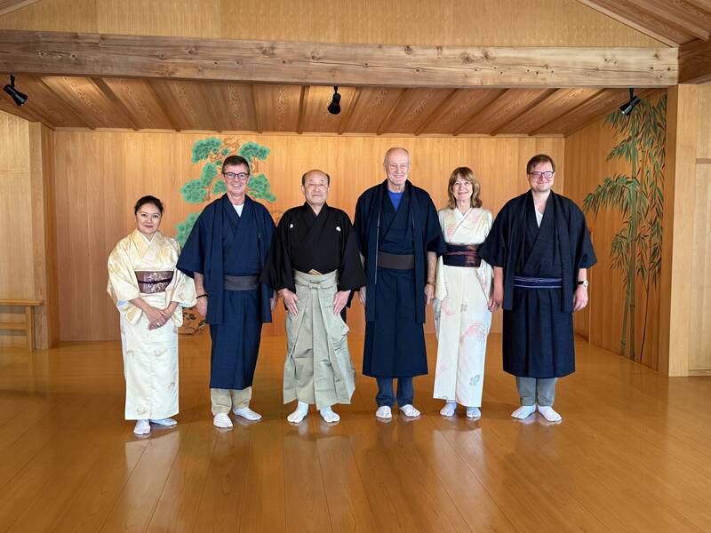 Sechs Personen in traditioneller japanischer Kleidung stehen lächelnd in einer hölzernen Halle vor einer bemalten Kulisse mit einem Baum.