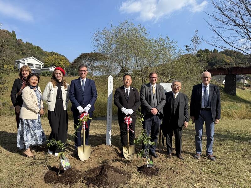 Acht Personen stehen in formeller Kleidung lächelnd auf einer Wiese neben frisch gepflanzten Bäumen mit Schaufeln im Boden.