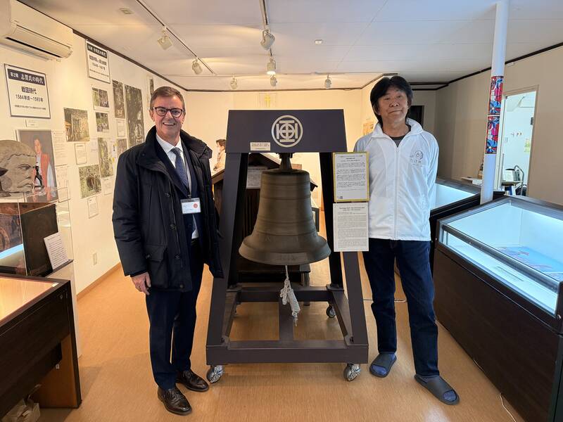Zwei Männer stehen in einem Ausstellungsraum neben einer großen Glocke auf einem Ständer.