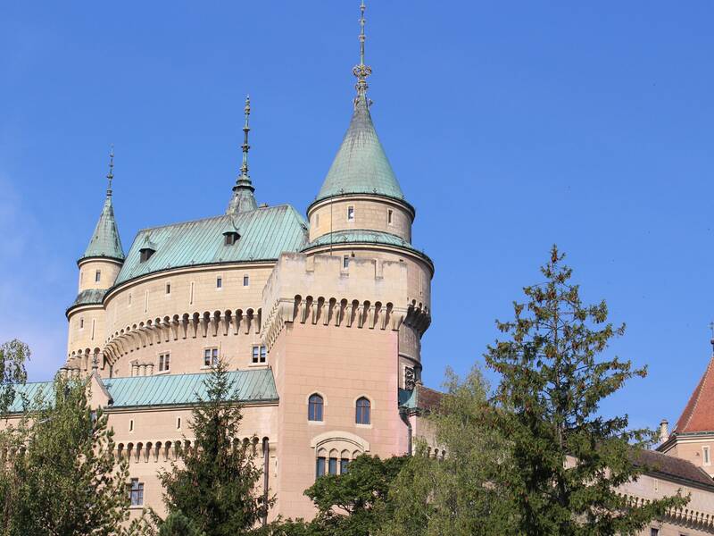 Eine Burg mit mehreren Türmen und grünlichen Dächern ragt hinter Bäumen vor einem klaren blauen Himmel auf. Bild aus unserer Partnerstadt Bojnice.