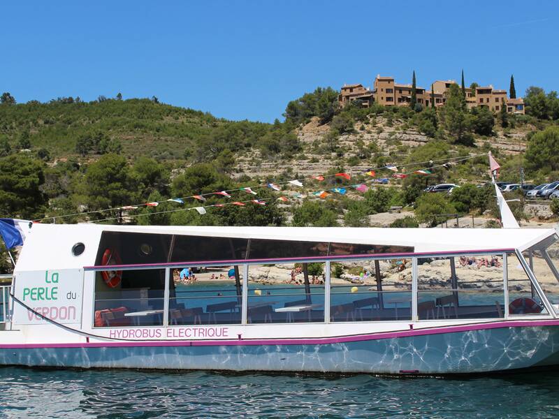 Ein elektrisches Passagierboot mit dem Namen "La Perle du Verdon" fährt auf blauem Wasser, umgeben von grünen Hügeln und einem Gebäude im Hintergrund.