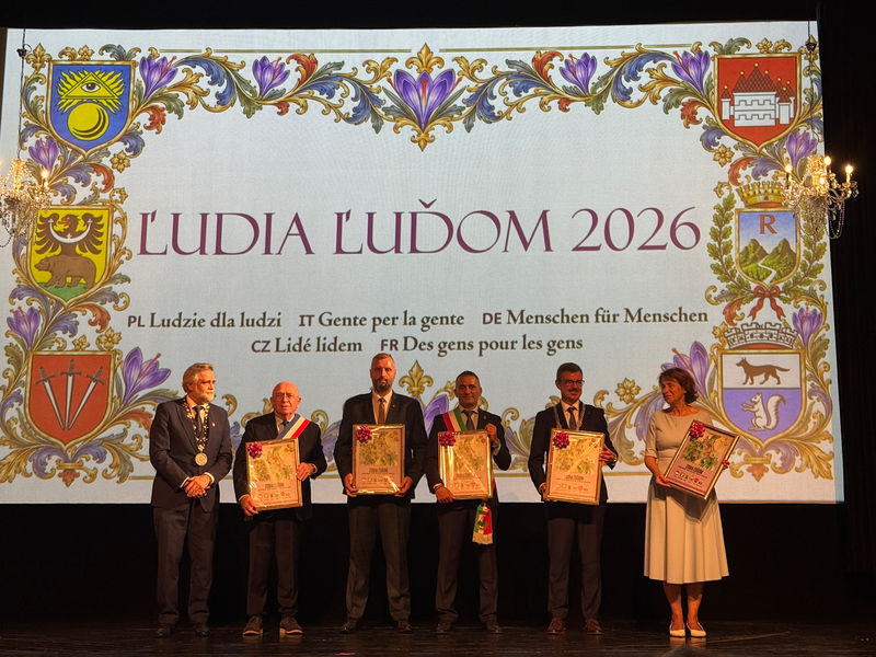 Fünf Personen stehen auf einer Bühne vor einem dekorativen Banner mit der Aufschrift "LUDIA LUĎOM 2026" und halten gerahmte Urkunden in den Händen.