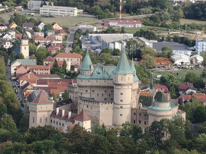 Ein großes, historisches Schloss mit mehreren Türmen und grünen Dächern ist von einer Stadt und viel Grün umgeben.