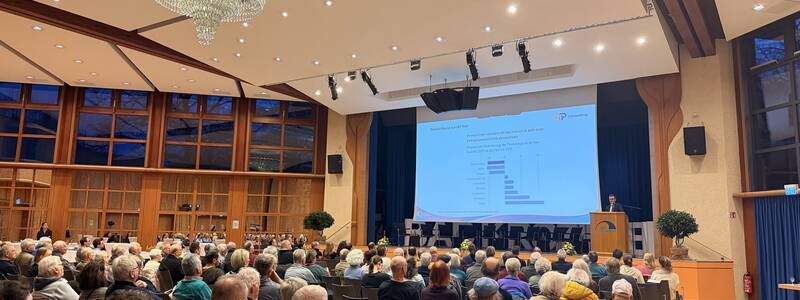 Ein großer, gut besuchter Veranstaltungsraum mit einem Redner, der an einem Pult vor einer Leinwand mit einer Präsentation steht, während das Publikum aufmerksam zuhört.