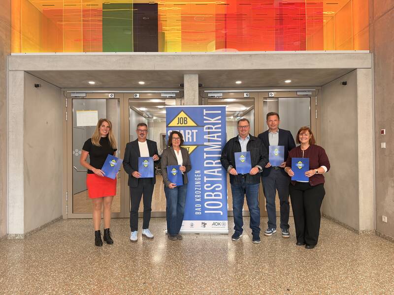 Eine Gruppe von sechs Personen steht vor einem Eingang und hält blaue Mappen, mit einem Plakat im Hintergrund, das mit "Job Start Markt" beschriftet ist.