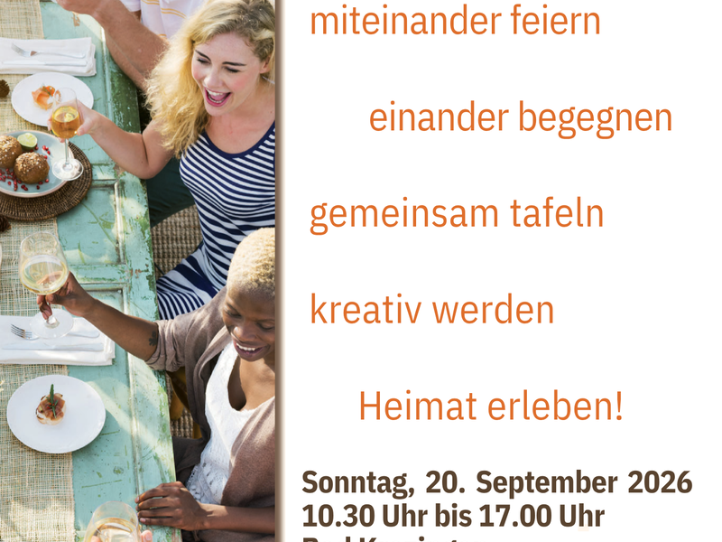 Eine Gruppe von Menschen sitzt lachend an einem gedeckten Tisch bei einem Fest mit dem Motto "Zusammen sind wir Heimat".